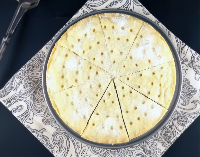 Overhead shot of a round shortbread in a pan cut into slices with holes on the top and a sprinkling of white sugar on a paisley cloth