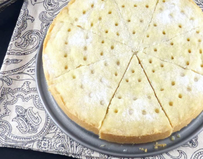 Side view shot of a round shortbread in a pan cut into slices with holes on the top and a sprinkling of white sugar on a paisley cloth