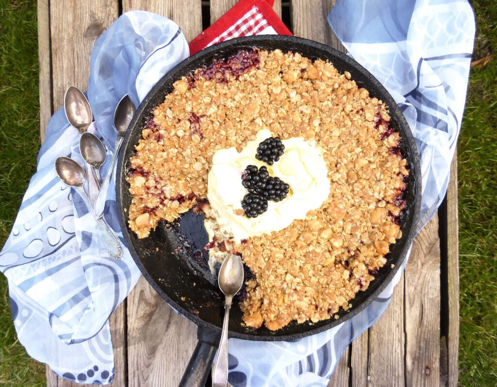 picture of an apple and blackberry dessert in a skillet with a blue scarf and tablespoons by the side