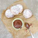four doughnuts with a small bowl of Nutella and a spoon on a sheet of brown paper