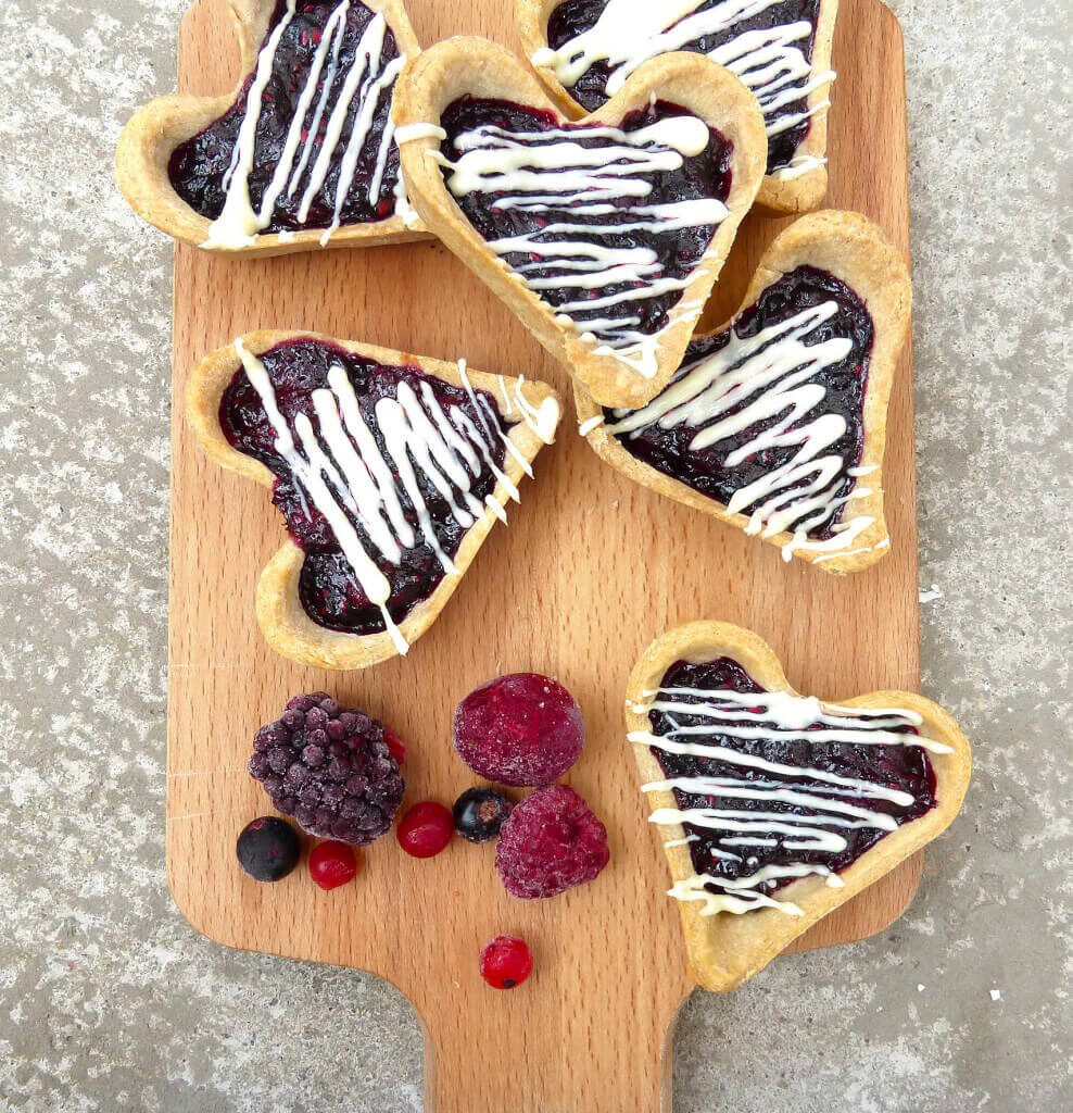 homemade heart shaped jam tarts on a wooden chopping board with mixed berries by the side