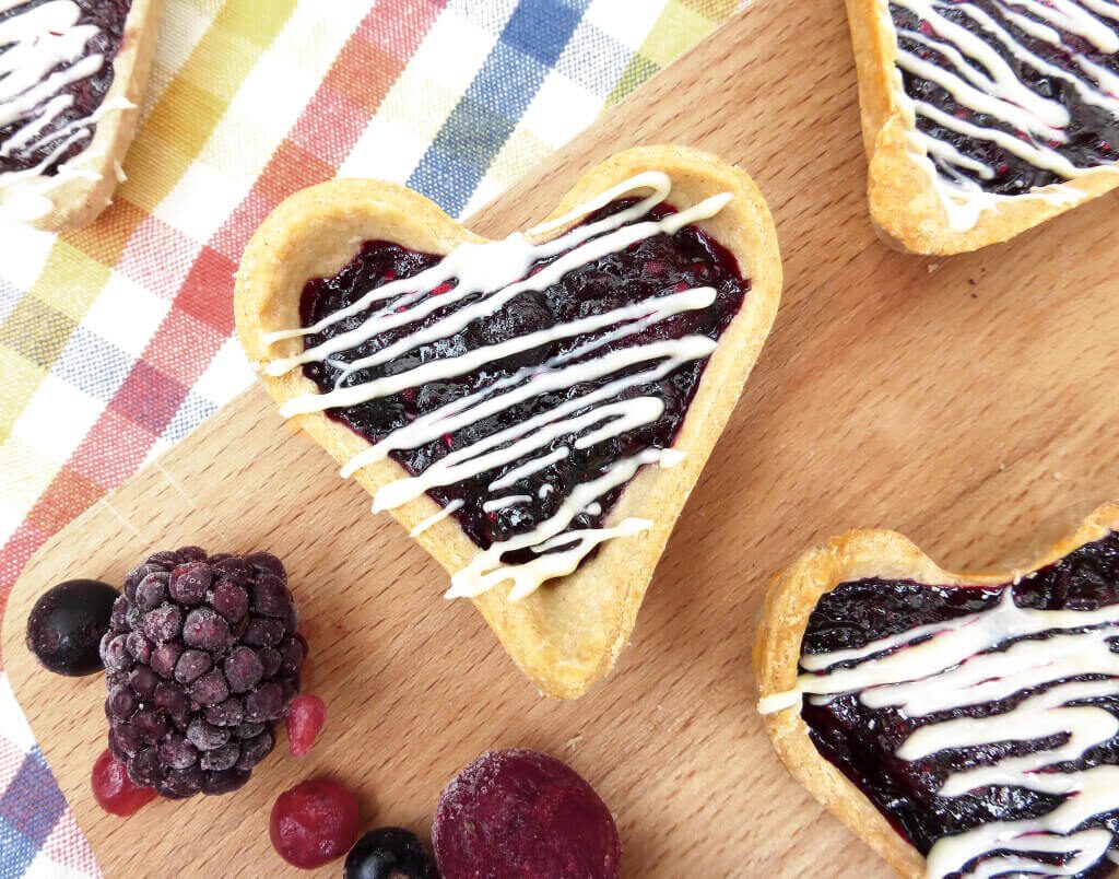 picture of a jam tart drizzled with white chocolate on a wooden board with a blackberry and raspberry next to it