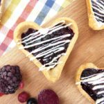 picture of a jam tart drizzled with white chocolate on a wooden board with a blackberry and raspberry next to it