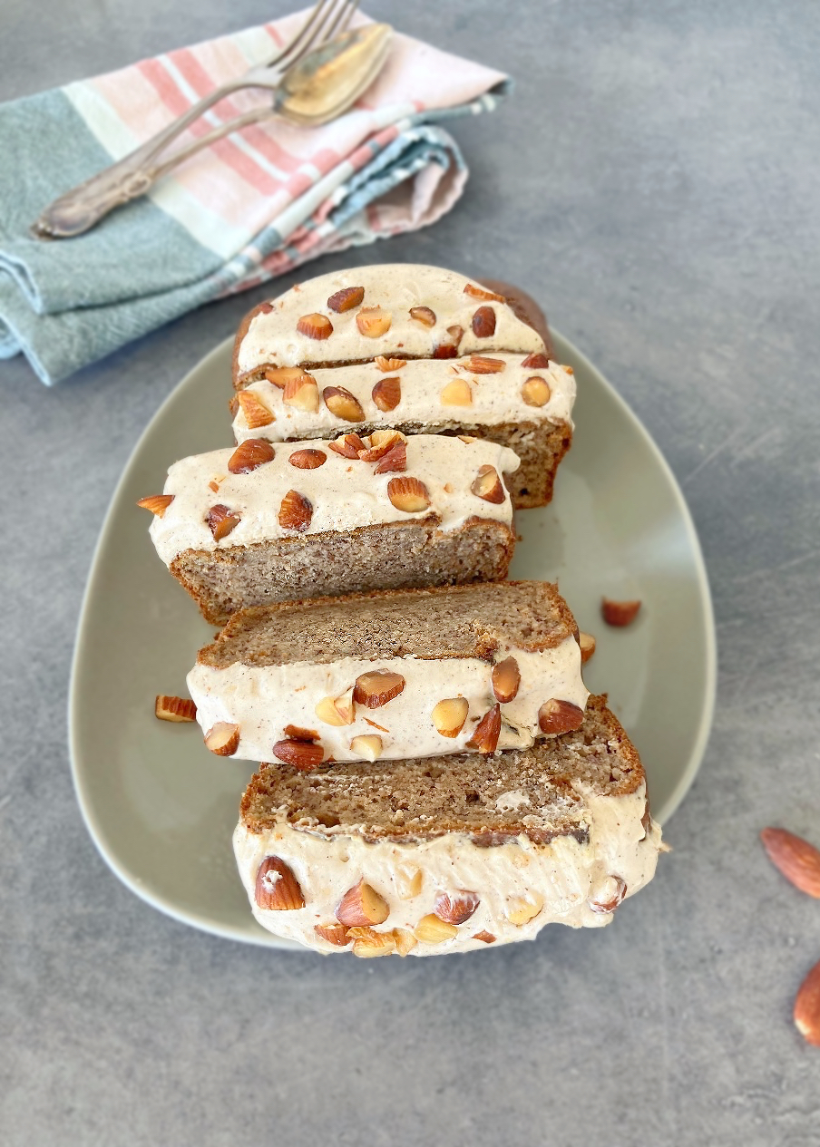 Banana bread loaf on a green plate, sliced into thick slices and topped with maple syrup cream cheese frosting and a sprinkling of almonds.