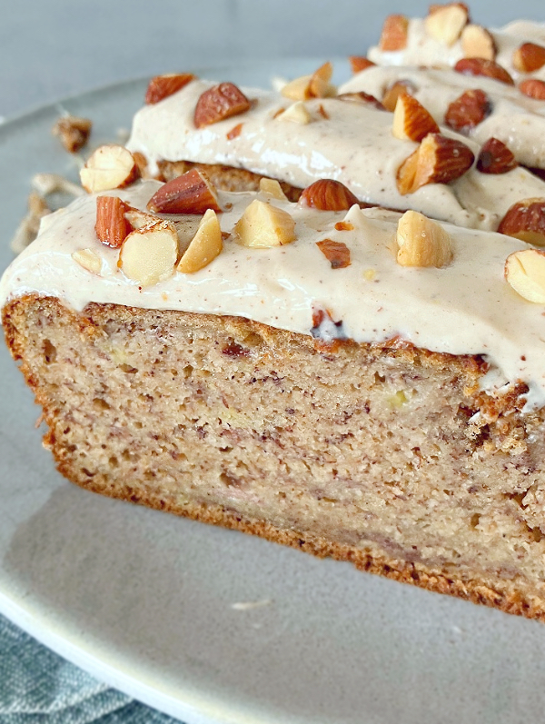 Close up of a slice of banana bread loaf on a green plate, and topped with maple syrup cream cheese frosting and a sprinkling of almonds.
