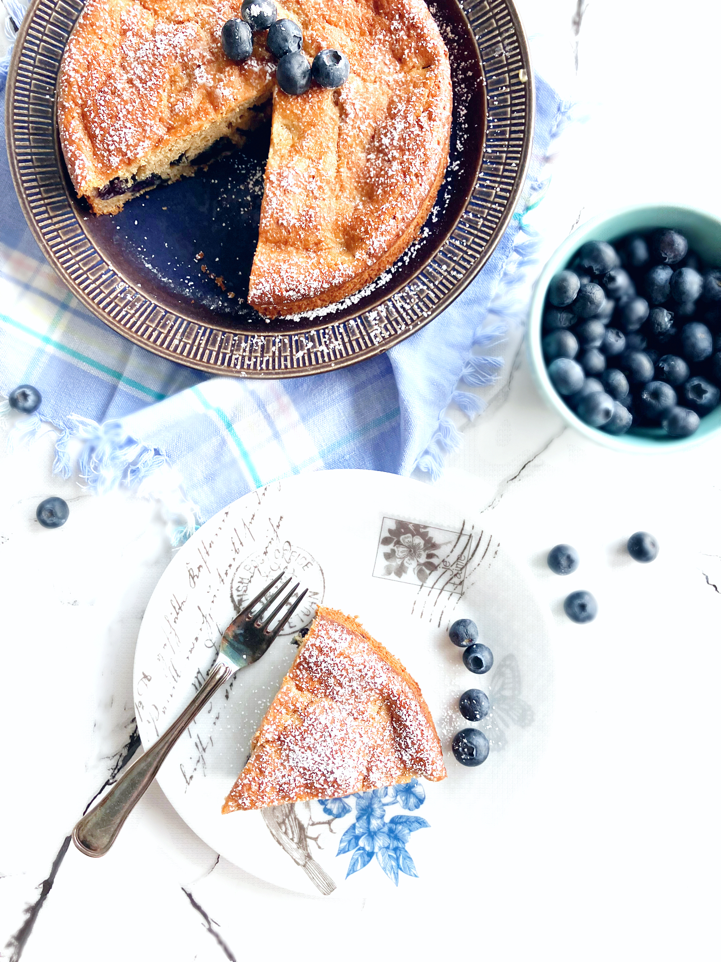 overhead shot of a blueberry and lime spelt cake with a slice cut out and a bowl of blueberries