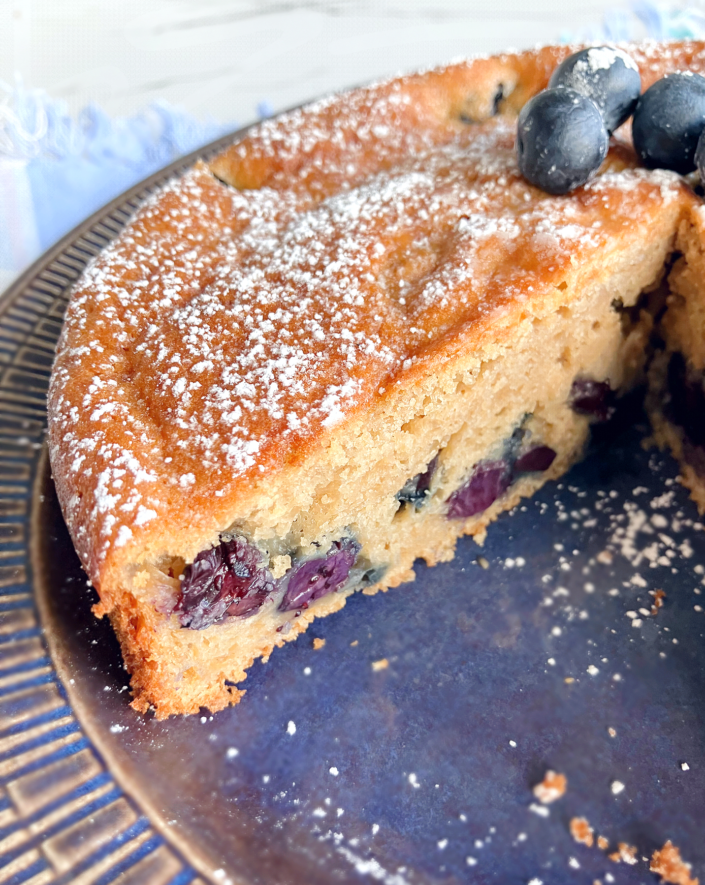 cut open blueberry and lime spelt cake showing blueberries in the middle, on a blue plate