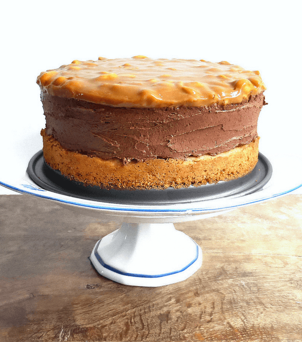 Decadent chocolate cheesecake with a peanut butter crust topped with salted peanut dulce de leche on a white stand on a wooden table