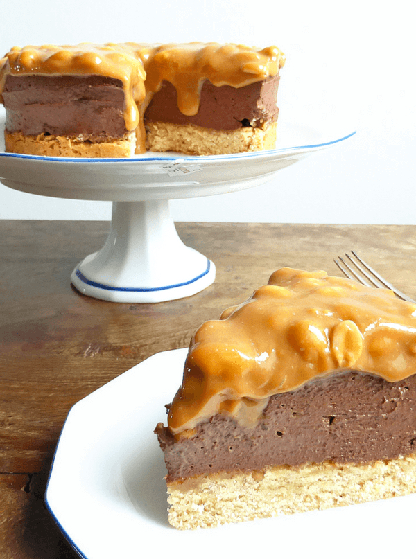 Decadent chocolate cheesecake with a peanut butter crust topped with salted peanut dulce de leche on a white stand on a wooden table