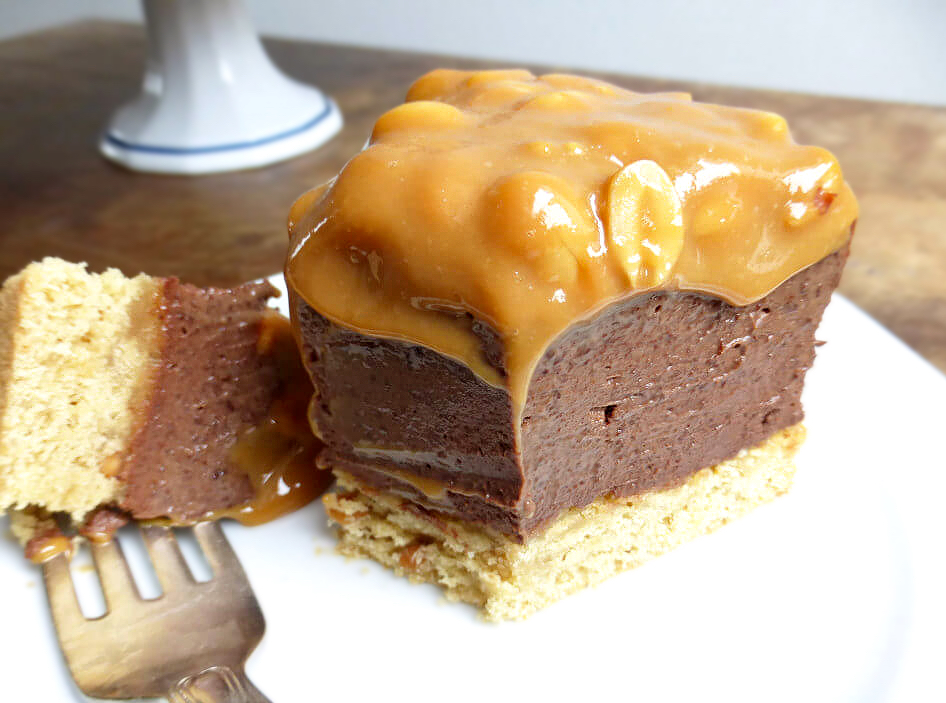 Slice of decadent chocolate cheesecake with a peanut butter crust topped with salted peanut dulce de leche on a white stand on a wooden table