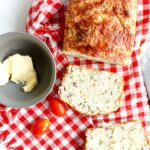 overhead view of cheese quick bread with two slices, butter and tomatoes on a checked red and white cloth