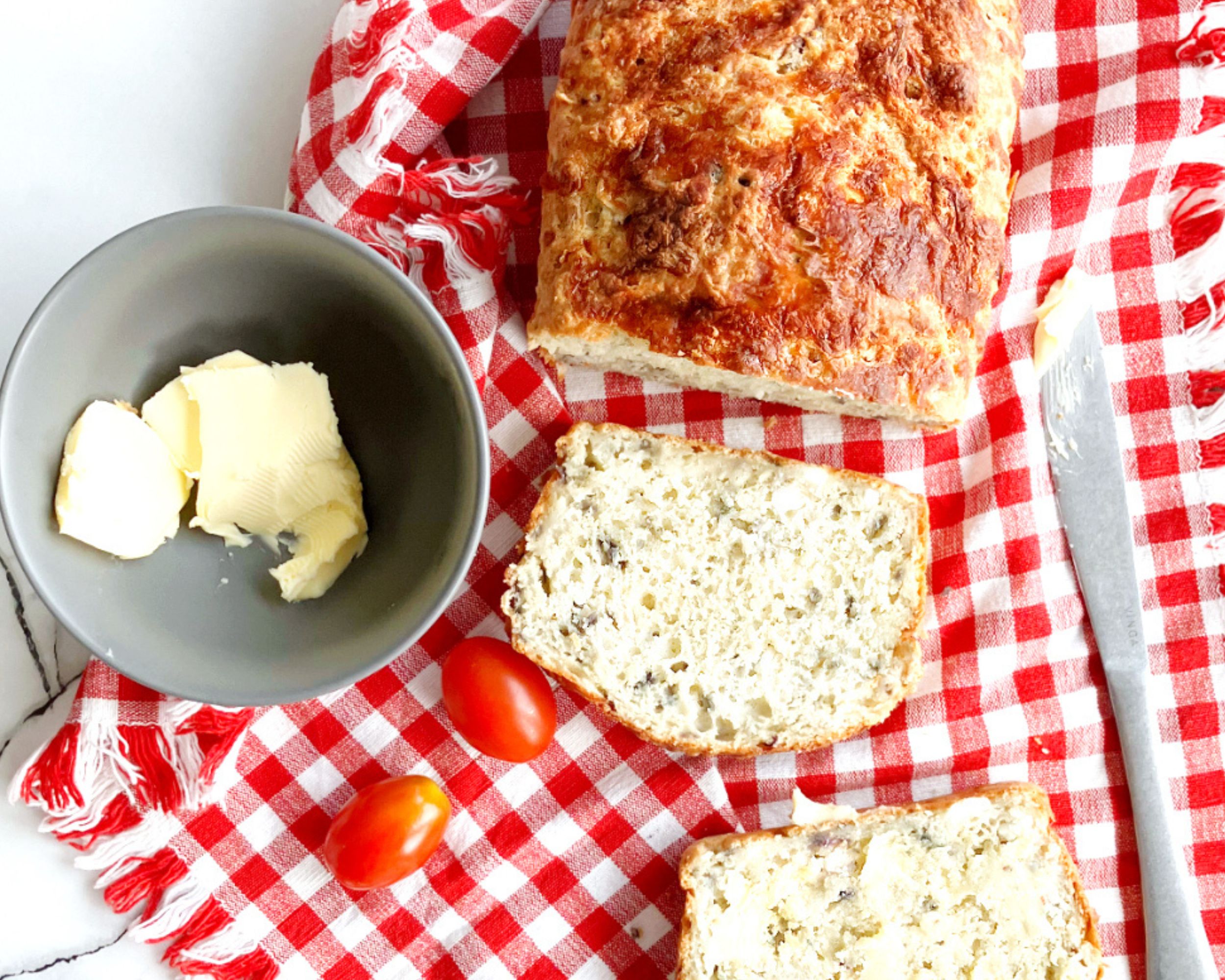 overhead view of cheese quick bread with two slices, butter and tomatoes on a checked red and white cloth
