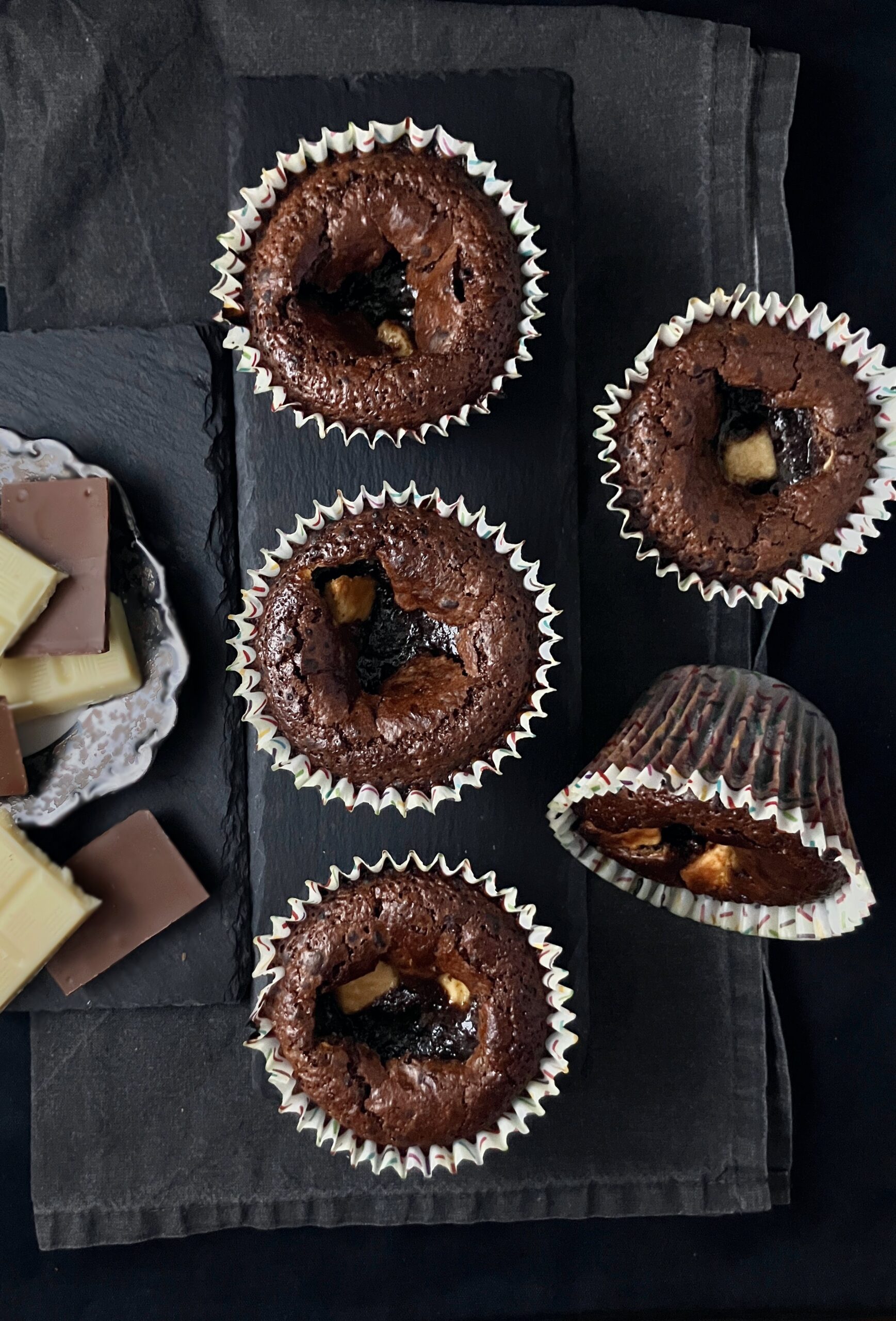 Five chocolate muffins on a dark background with white and dark chocolate to the side