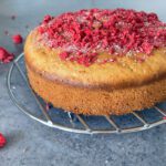 Side view shot of a cardamom and raspberry cake and freeze dried raspberries sprinkled around on a blue-grey surface