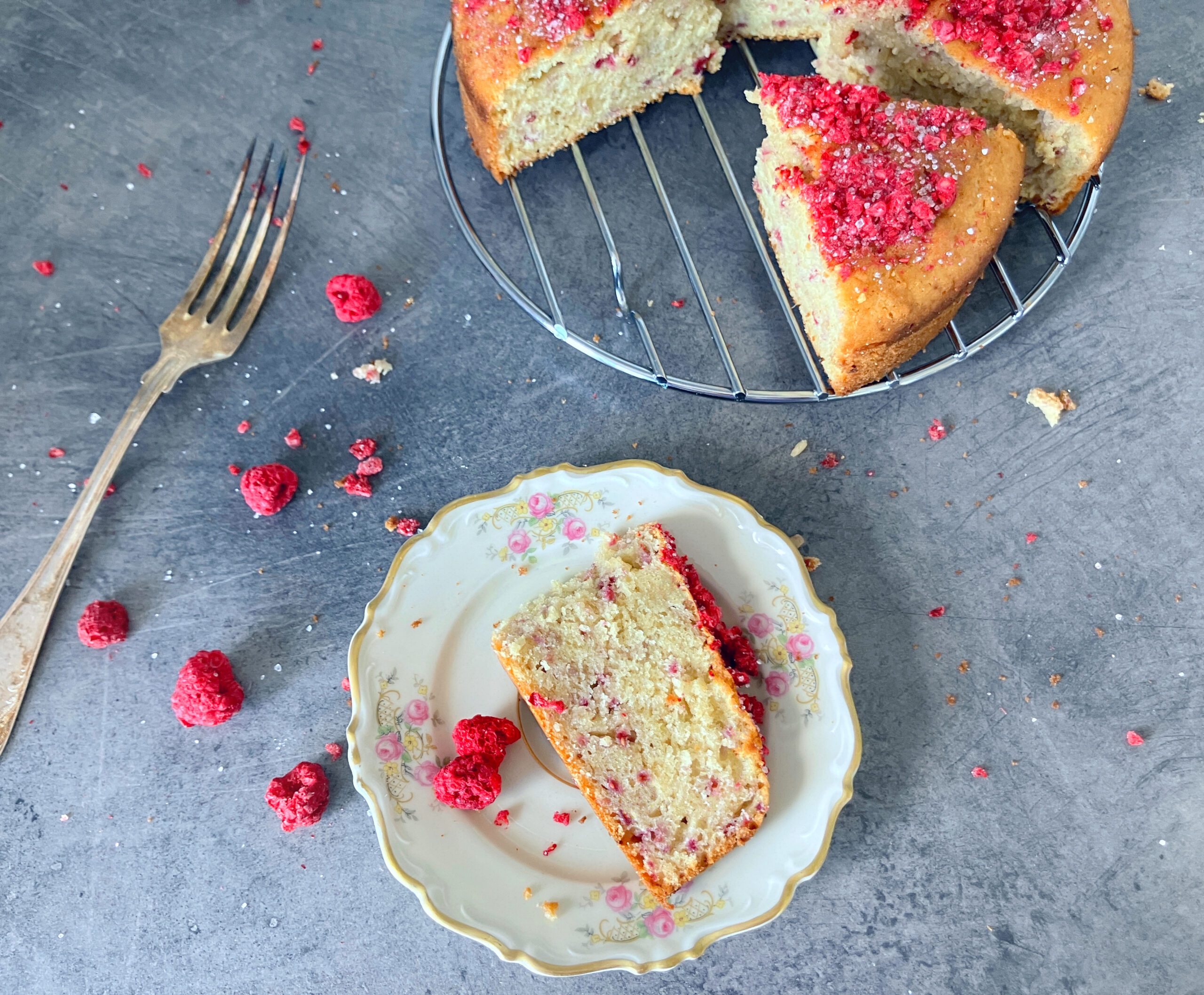 Overhead shot of a cardamom and raspberry cake with a slice missing and freeze dried raspberries sprinkled around on a blue-grey surface