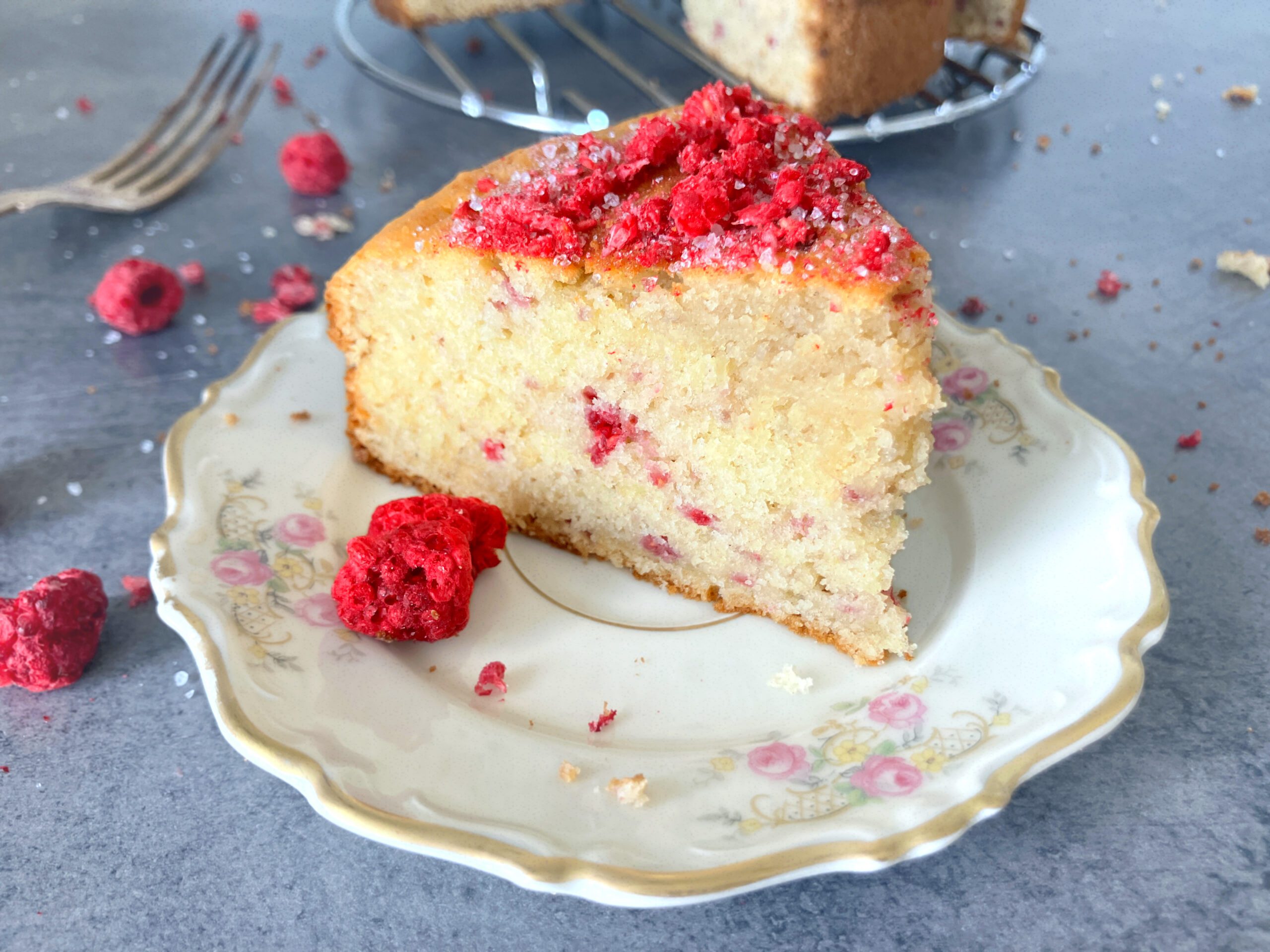 Close up shot of a slice of cardamom and raspberry cake and freeze dried raspberries sprinkled around on a blue-grey surface