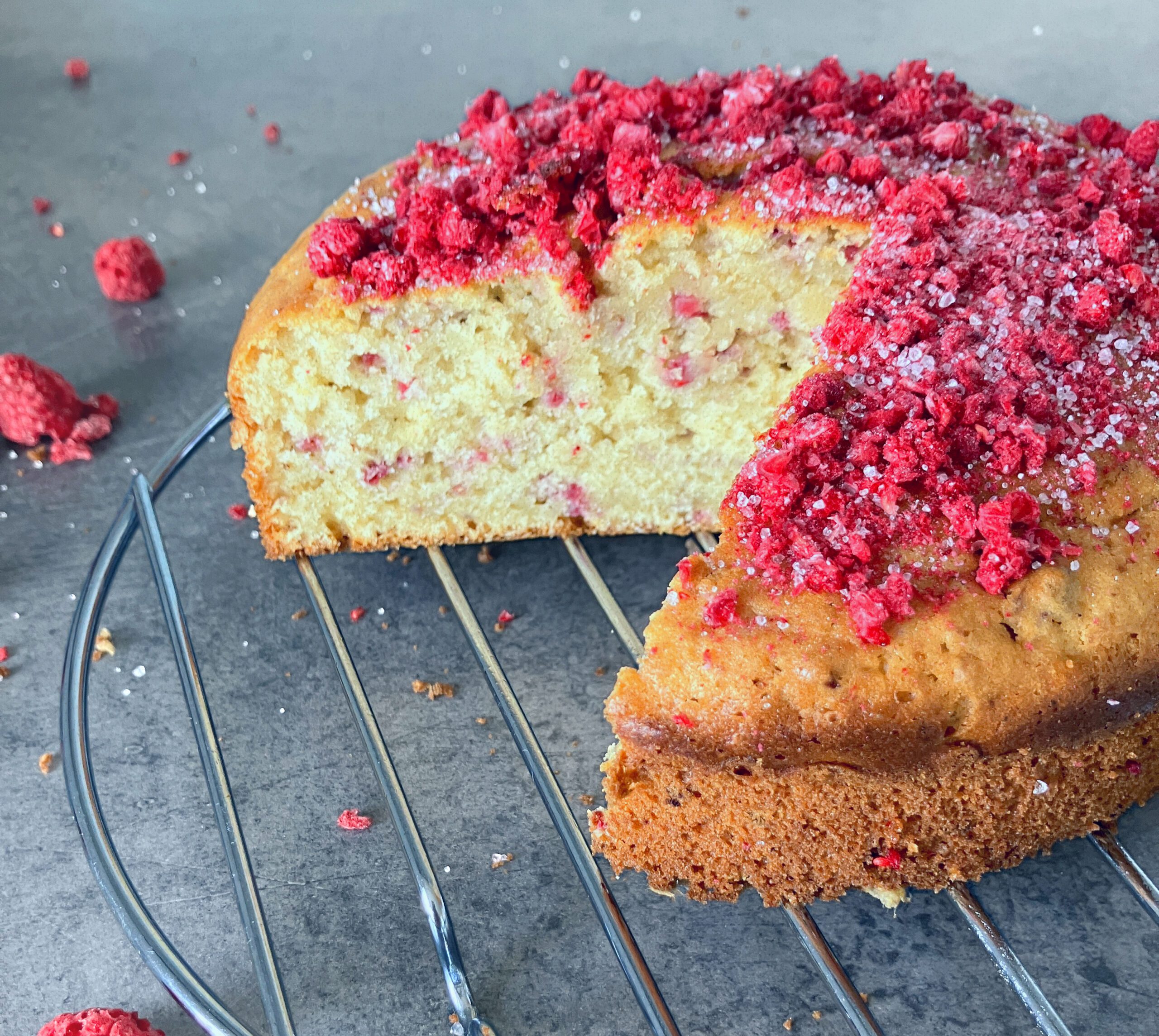 Close up shot of a slice of cardamom and raspberry cake and freeze dried raspberries sprinkled around on a blue-grey surface