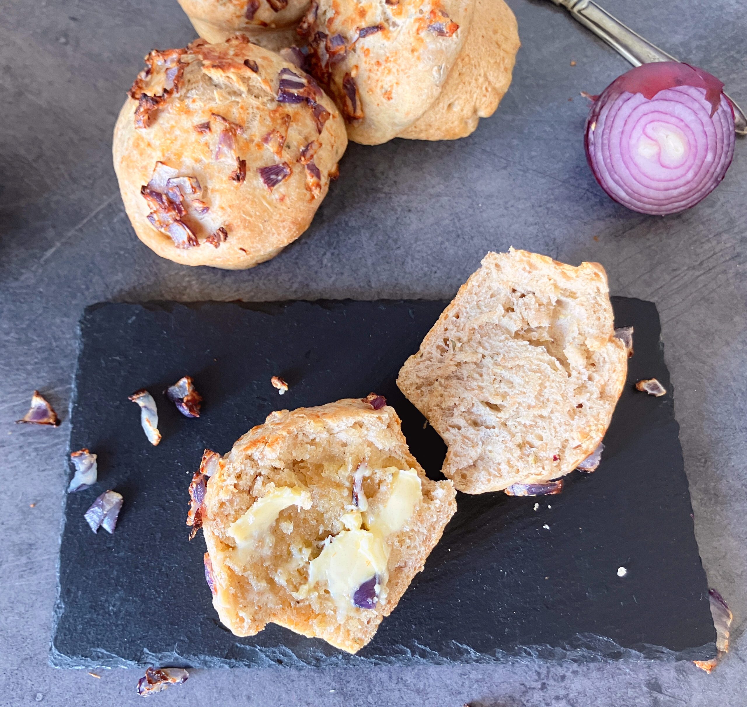 Spelt wholegrain buns with one cut in half with butter on, on a slate board with a half red onion behind