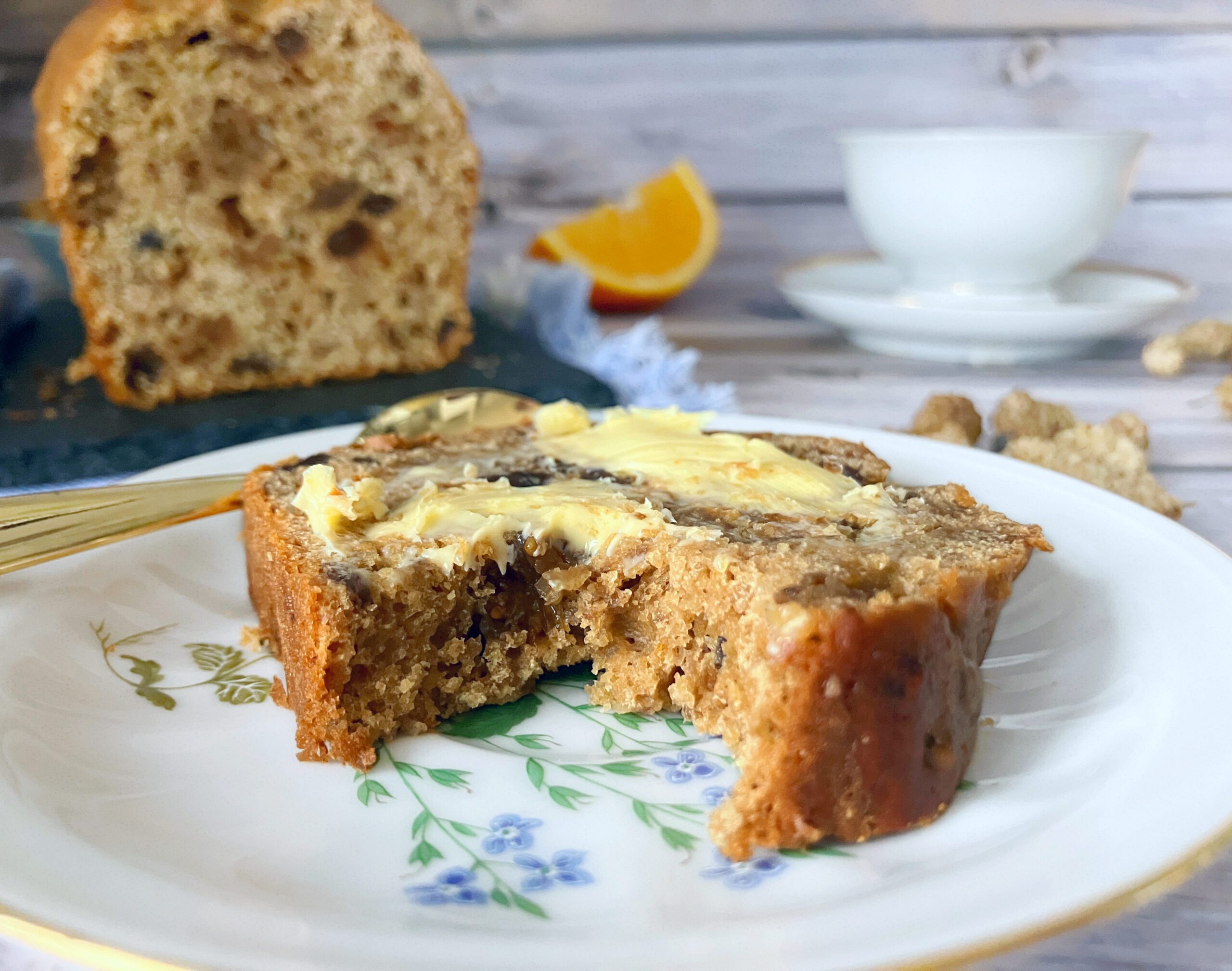 Dried mulberry tea loaf with a bite taken out, on a plate with a cup of tea and oranges